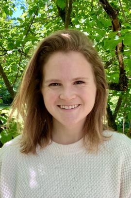 woman with light brown hair and a light sweater with greenery in the background